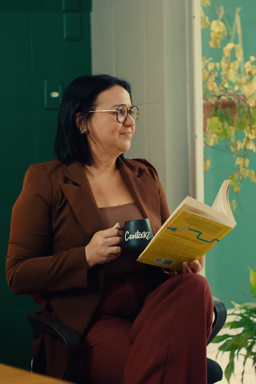 Ketlyn lendo com a caneca da ContaKE nas mãos, junto à janela do escritório